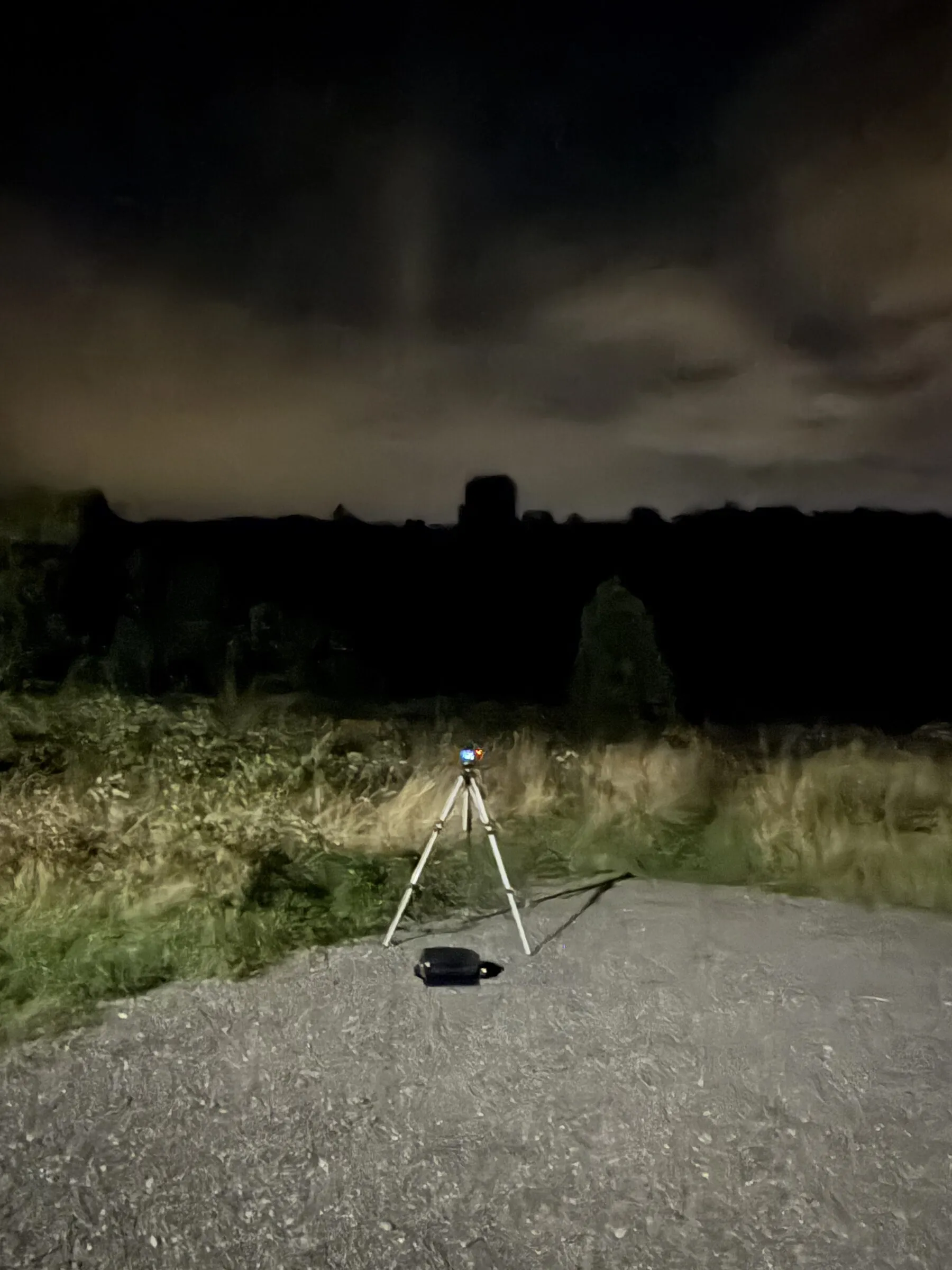 A camera on a small tripod on a gravel path at night, in front of the dark silhouette of Landskron castle.